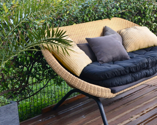 Wicker outdoor bench with black cushion and assorted pillows on wooden deck